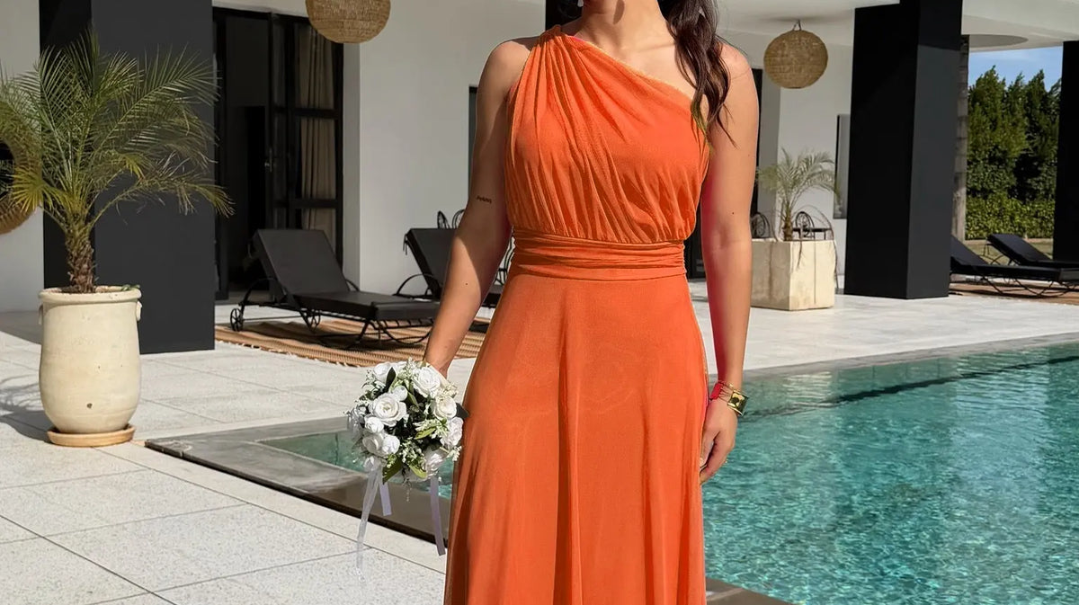 Robe longue orange à encolure asymétrique et taille marquée, portée avec un bouquet au bord d’une piscine.