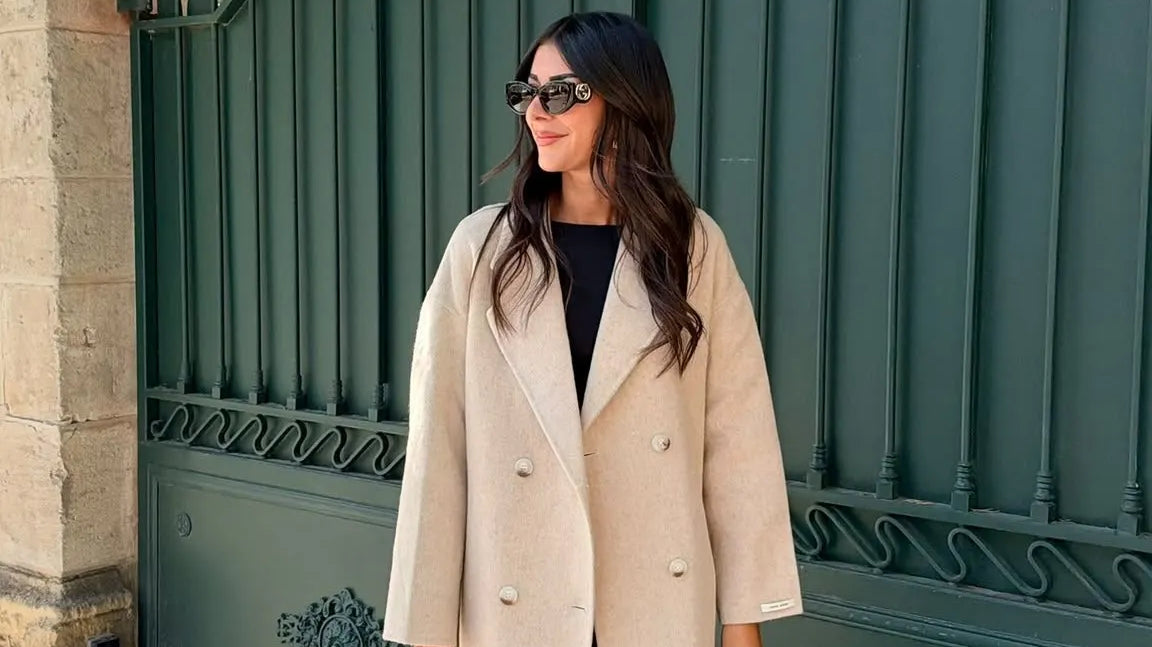 Trench long beige à double boutonnage, porté avec des bottines noires et un sac noir.