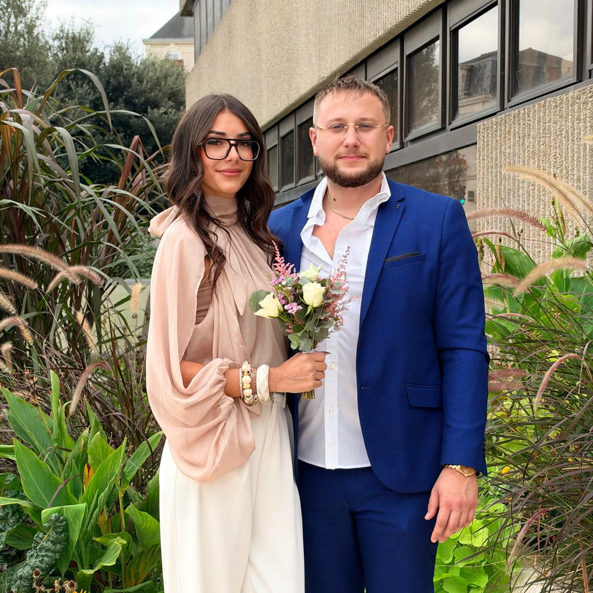Couple posant avec un bouquet de fleurs, la femme en tenue rose poudré et blanche et l’homme en costume bleu.