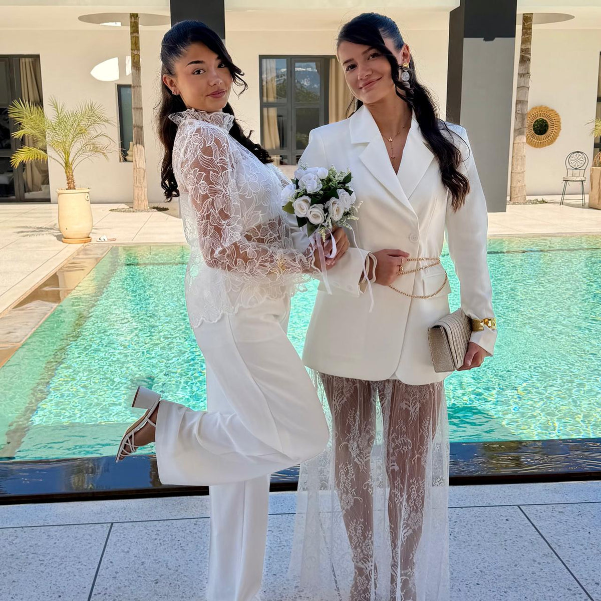 Deux femmes en tenues blanches de cérémonie, associant dentelle, pantalon et blazer au bord d’une piscine.