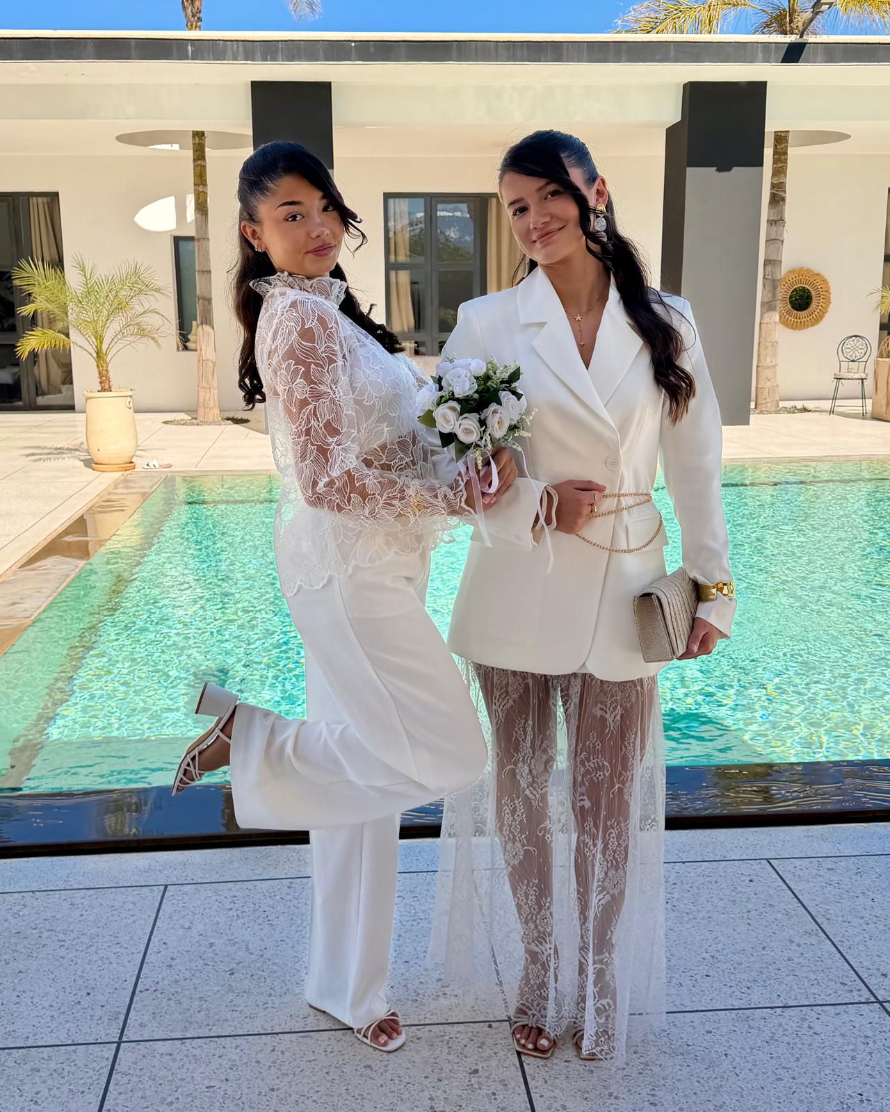 Deux femmes en tenues blanches de cérémonie, associant dentelle, pantalon et blazer au bord d’une piscine.